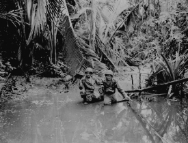 cameramen wading through stream while following infantry troops