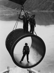 men riding on large casing section of pipe suspended by cable