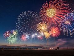 Brilliant bursts of fireworks cascade across the deep blue night sky, creating a vibrant display over a serene countryside landscape.