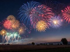 Vibrant fireworks burst in a colorful array above tranquil fields at dusk, creating a celebratory atmosphere that lights up the horizon with joy and brilliance.