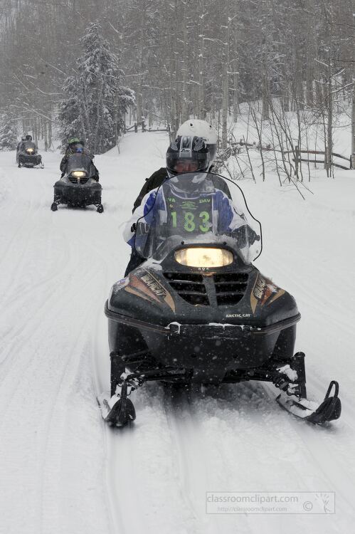  group of snowmobilers riding through a snowy forest trai