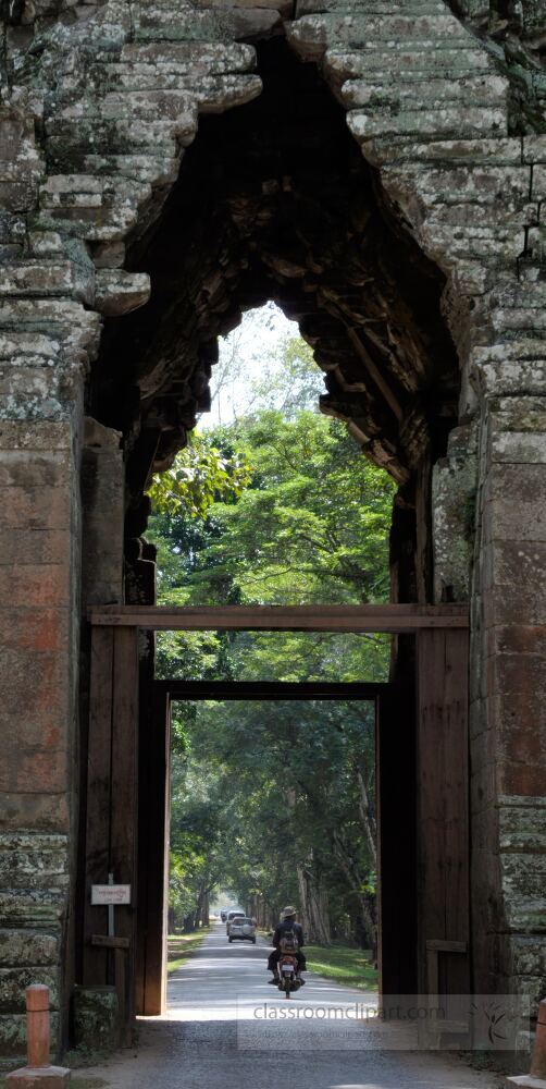 Bathed in natural light the ancient stone arch of Angkor Wat frames a serene path lined with lush greenery. A lone traveler rides through connecting history and present on this iconic route.