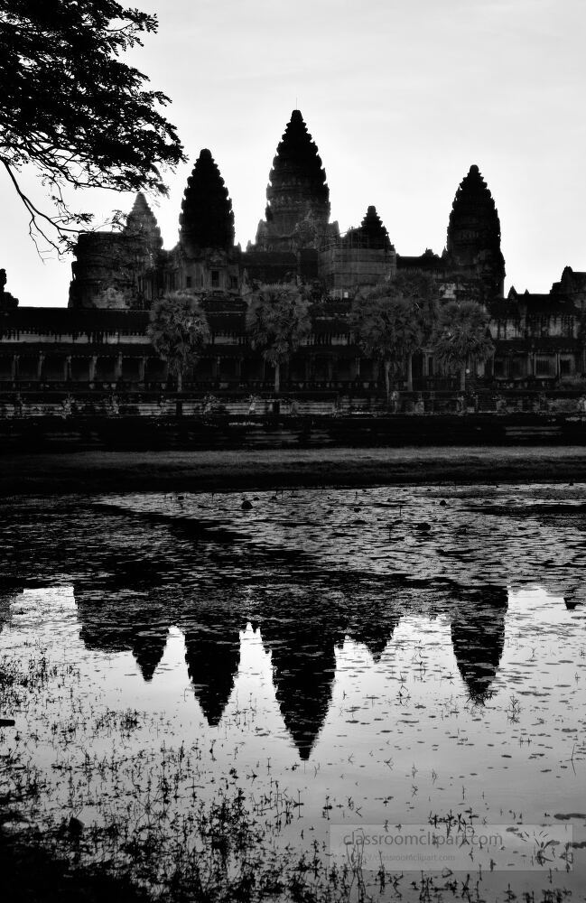 Towering spires of Angkor Wat emerge as shadows at dusk reflecting on the still waters of the surrounding landscape in Siem Reap creating a serene and mystical atmosphere.