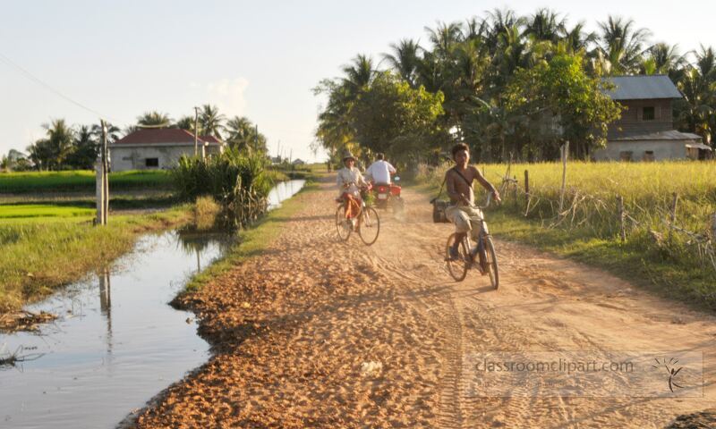 Bicycles glide along a serene dirt road in Siem Reap Cambodia. Surrounding lush greenery and tranquil waters evoke a peaceful rural atmosphere during the golden hour of the day.