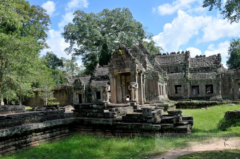 Majestic ruins of Angkor Wat stand against a backdrop of lush greenery and vibrant blue skies. Visitors wander through the enchanting site marveling at intricate stone carvings and historical significance.