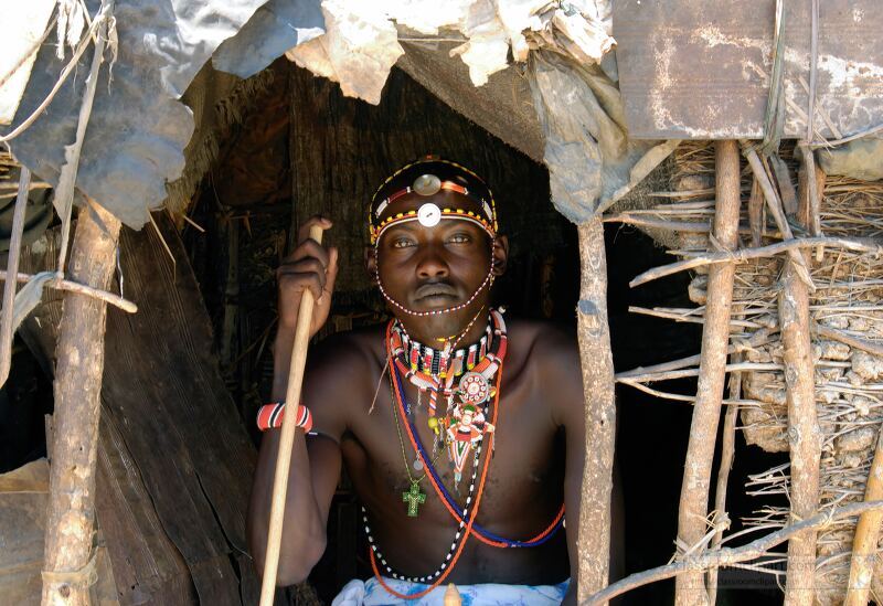 A Samburu tribesman sits at the entrance of his traditional dwelling in Kenya Adorned with colorful jewelry and cultural attire he holds a staff while showcasing the rich heritage of the Samburu people in their natural environment