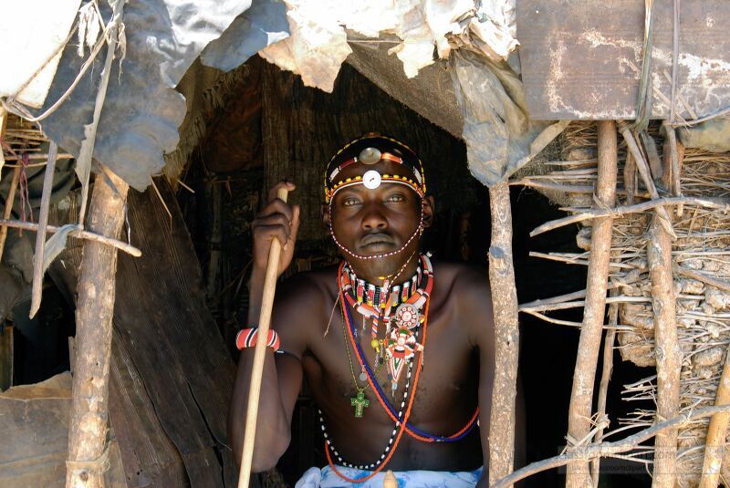 A Samburu tribesman dressed in vibrant traditional attire sits at the entrance of a mud hut Adorned with intricate beaded jewelry he holds a wooden staff showcasing the rich cultural heritage of the Samburu Tribe in Kenya