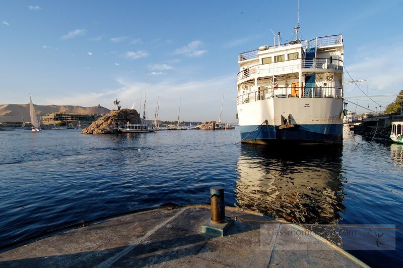 A large boat is docked on the calm waters of the Nile River Sailboats float nearby as the sun sets casting a warm glow over the scenic Aswan landscape