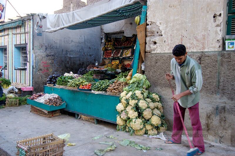 A fruit and vegetable vendor tends to his stall in Aswan Egypt Fresh produce is on display and the vendor is actively cleaning the ground showcasing the bustling market life