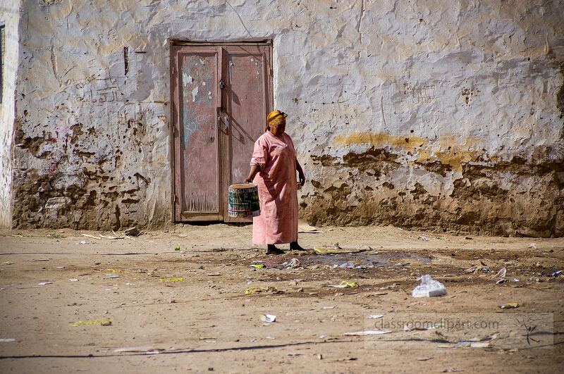 A woman in traditional clothing stands alone in a dusty area of Aswan Egypt holding a water container The background features a weathered wall and a closed door